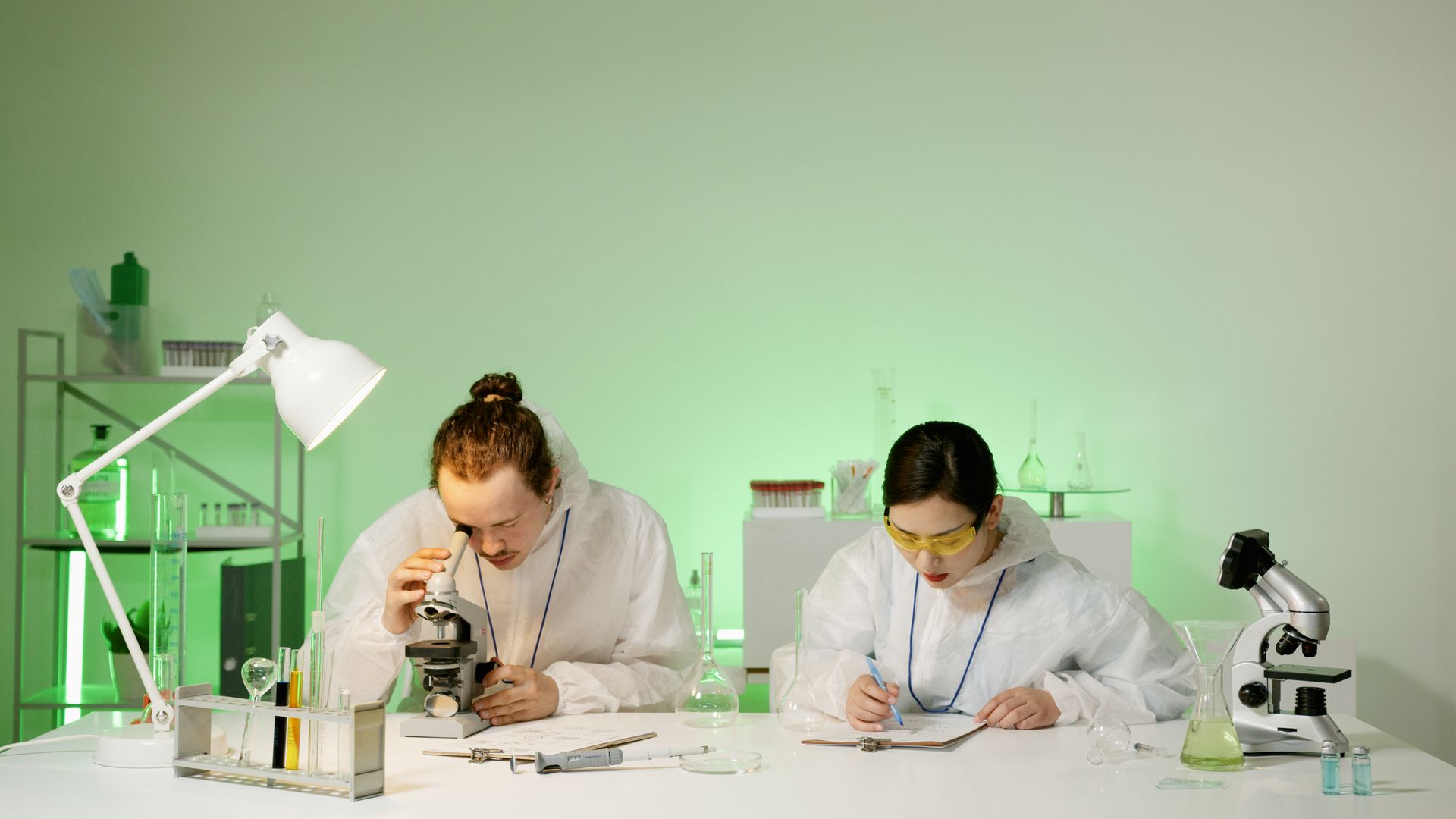 Two scientists in protective suits working with microscopes and notes in a laboratory setting.