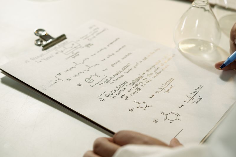 A close-up view of handwritten chemical formulas on a clipboard in a lab setting.