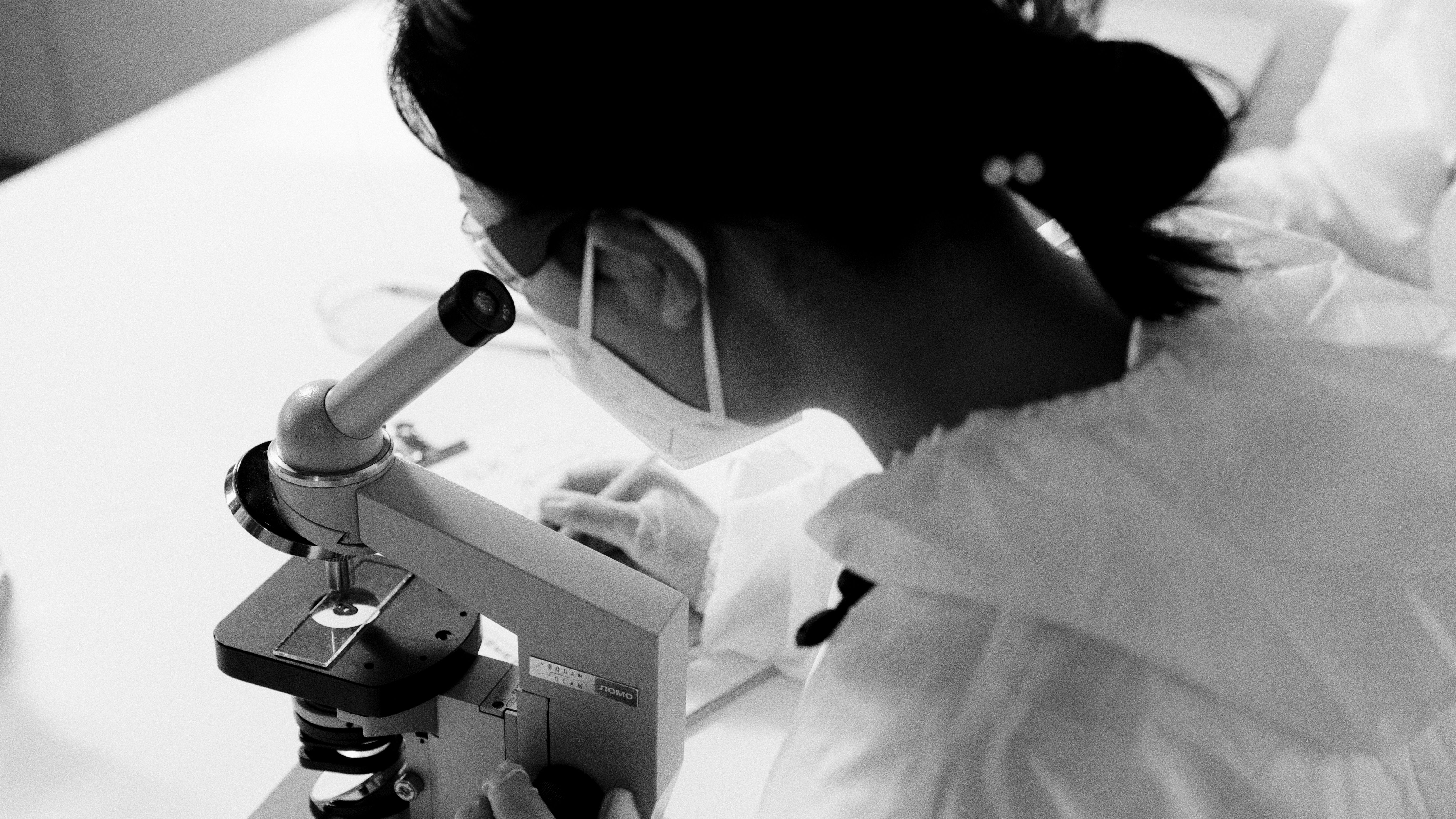 マスクと保護メガネを着用した科学者が顕微鏡で調査している白黒画像です。