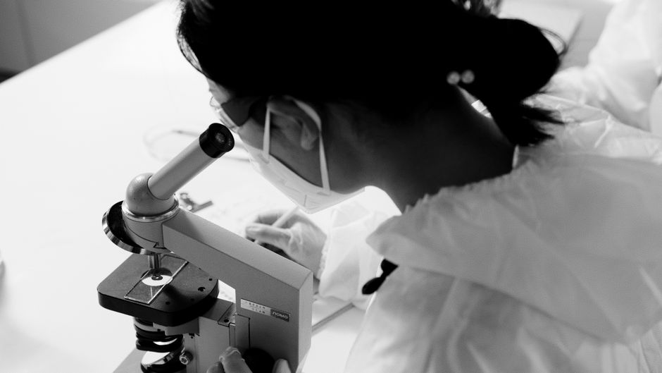 マスクと保護メガネを着用した科学者が顕微鏡で調査している白黒画像です。