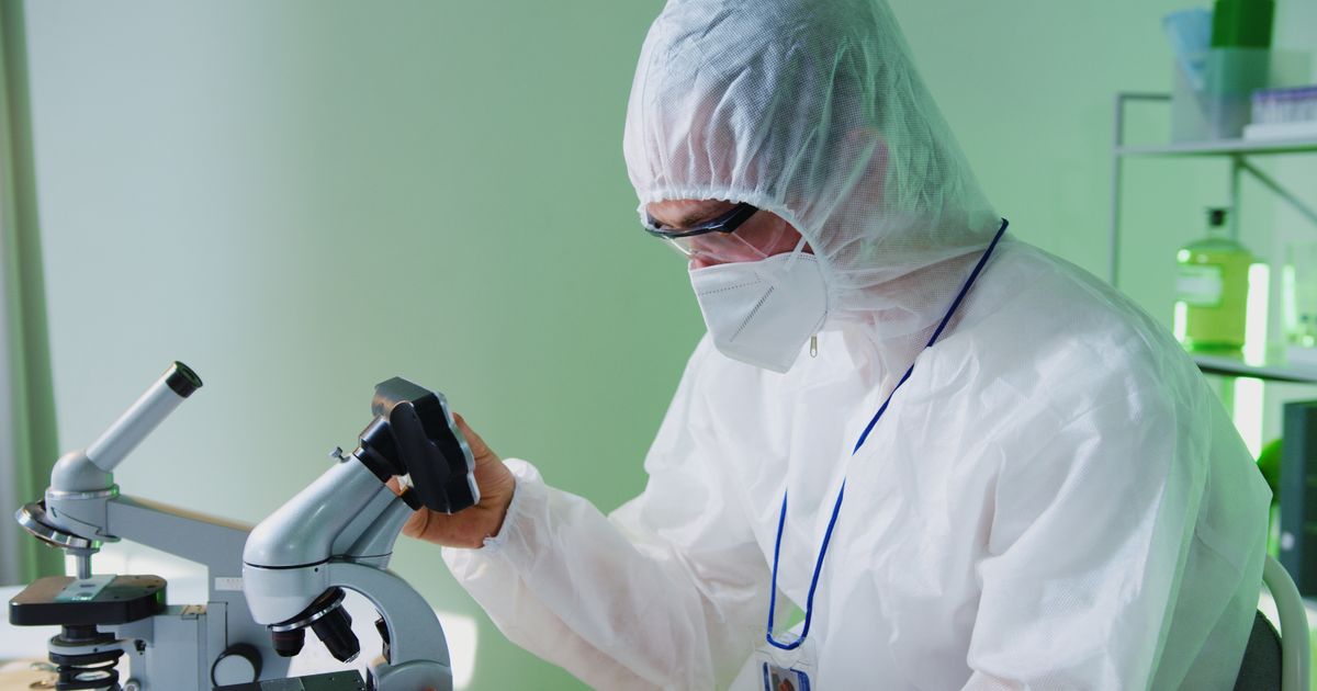 Scientist in protective gear examining samples under a microscope.