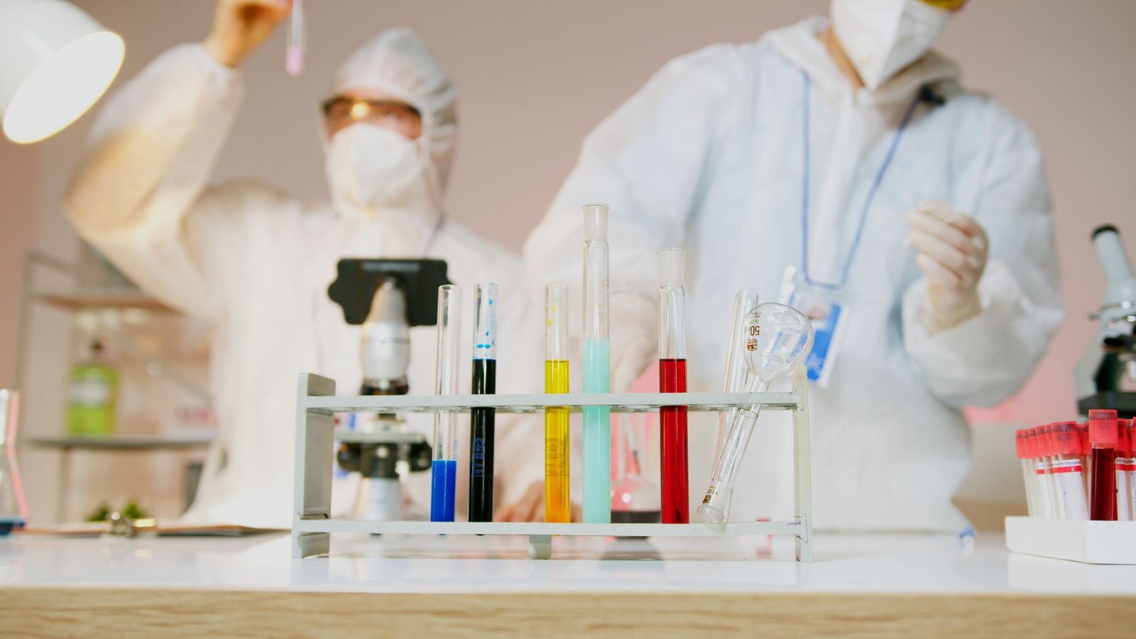 Two technicians in protective gear testing coloured liquid samples in a laboratory