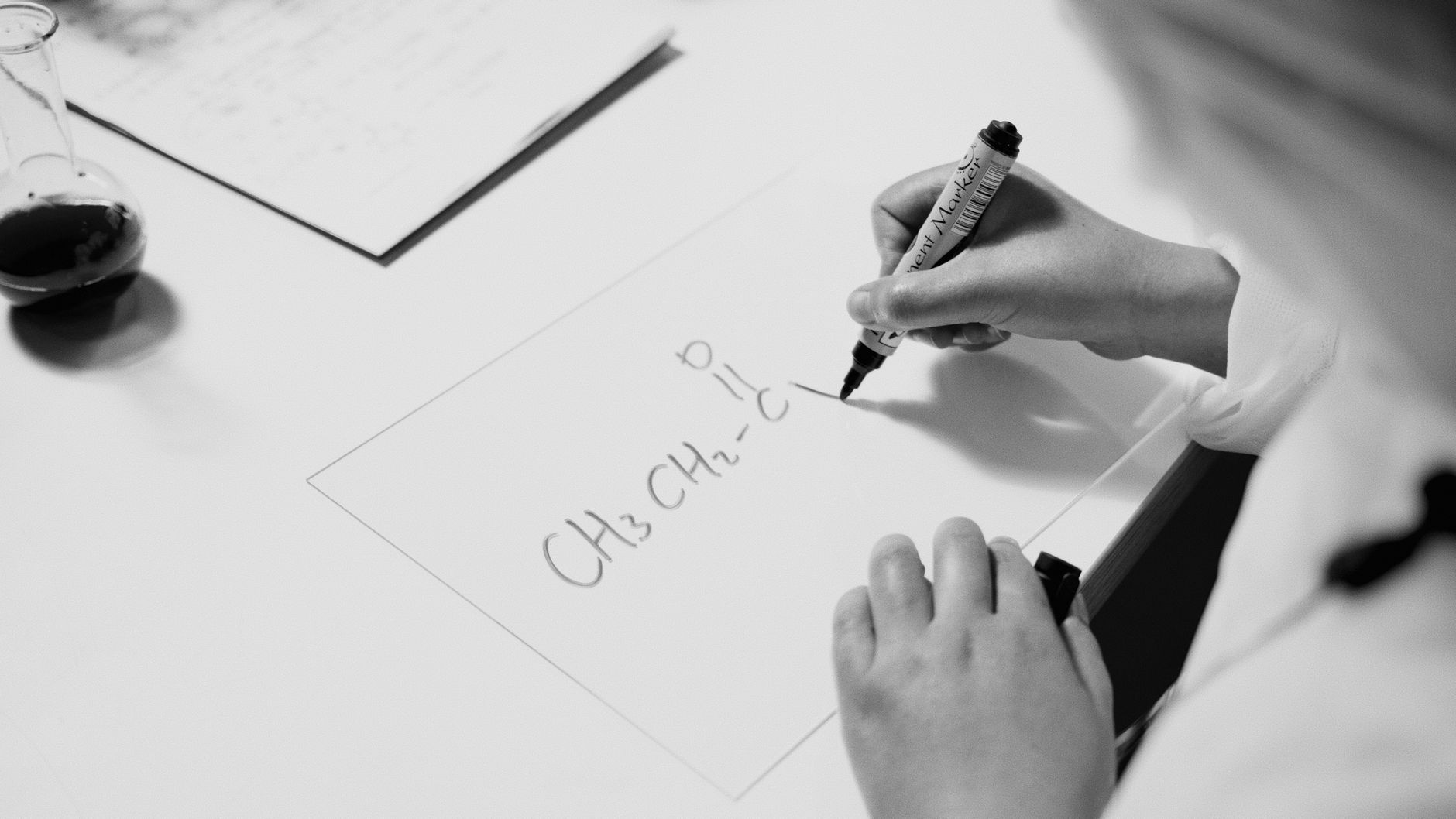 Black and white photo of hands writing a chemistry formula with a marker on a desk.