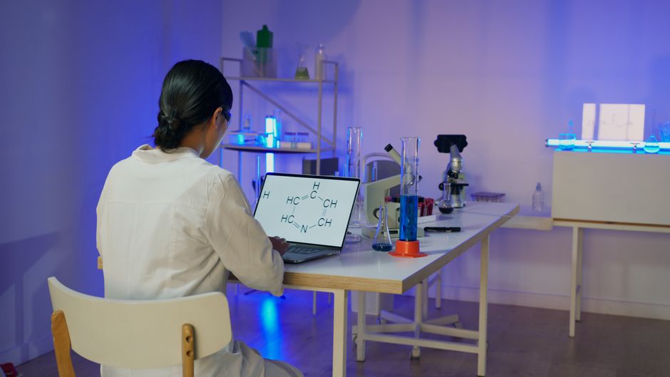 Woman in lab coat analyzing chemical formula on laptop in modern laboratory setting.