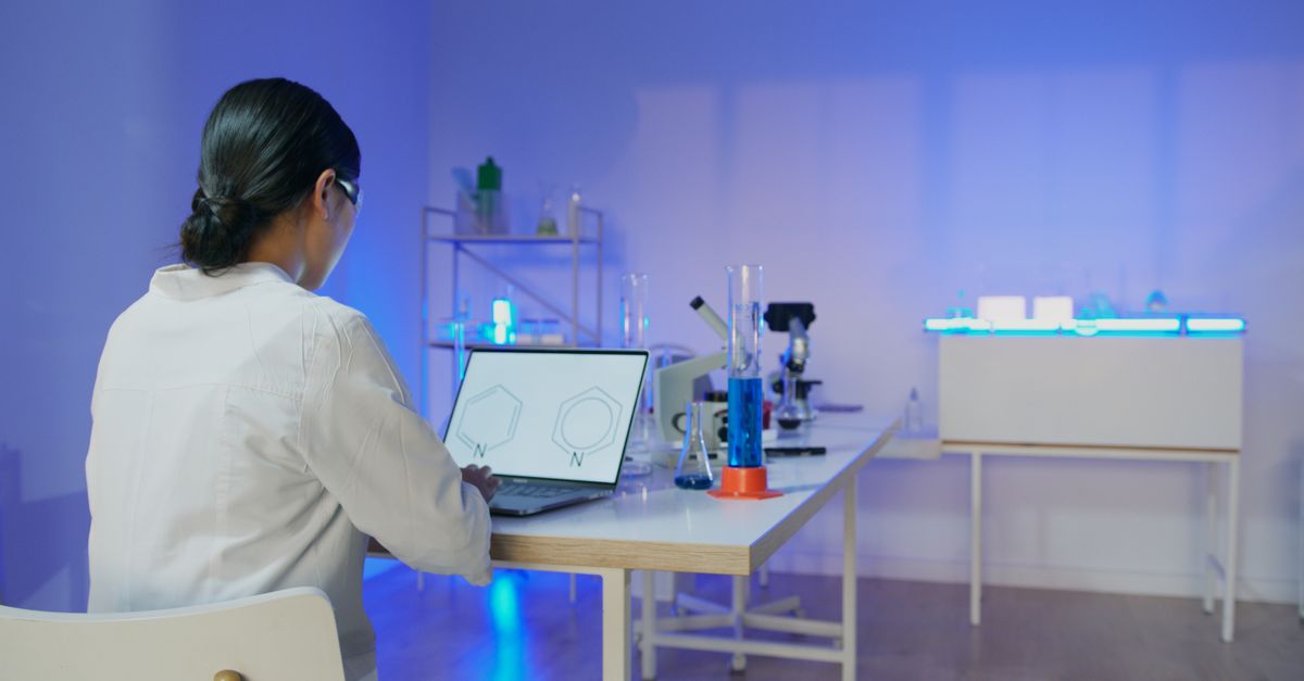 A scientist typing on a laptop in a lab, surrounded by beakers and glowing screens