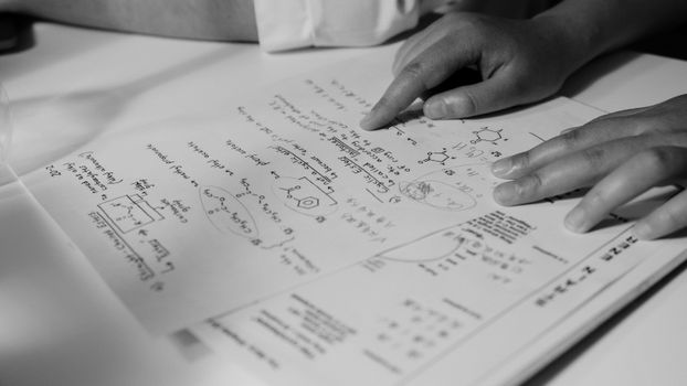 Grayscale photograph of hands pointing at handwritten scientific notes on a paper.
