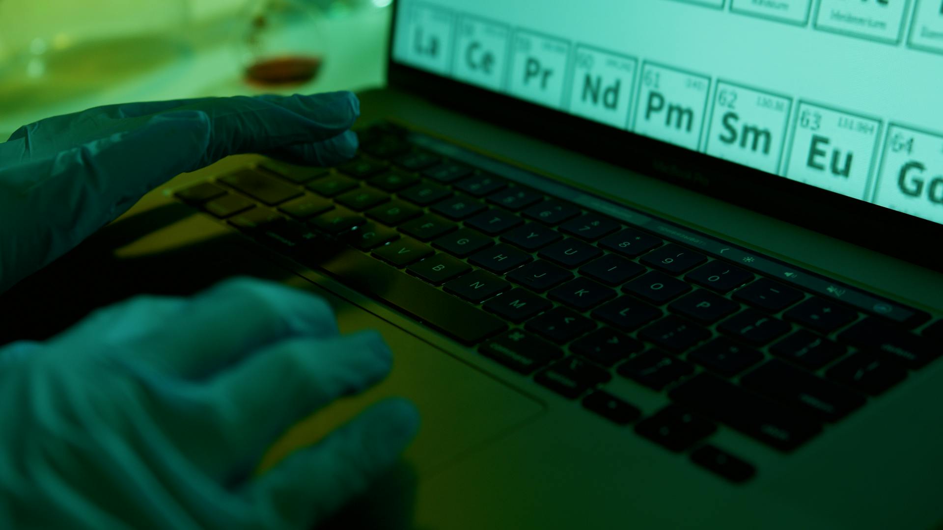 A scientist wearing gloves types data on a laptop in a laboratory setting, focused on the periodic table.