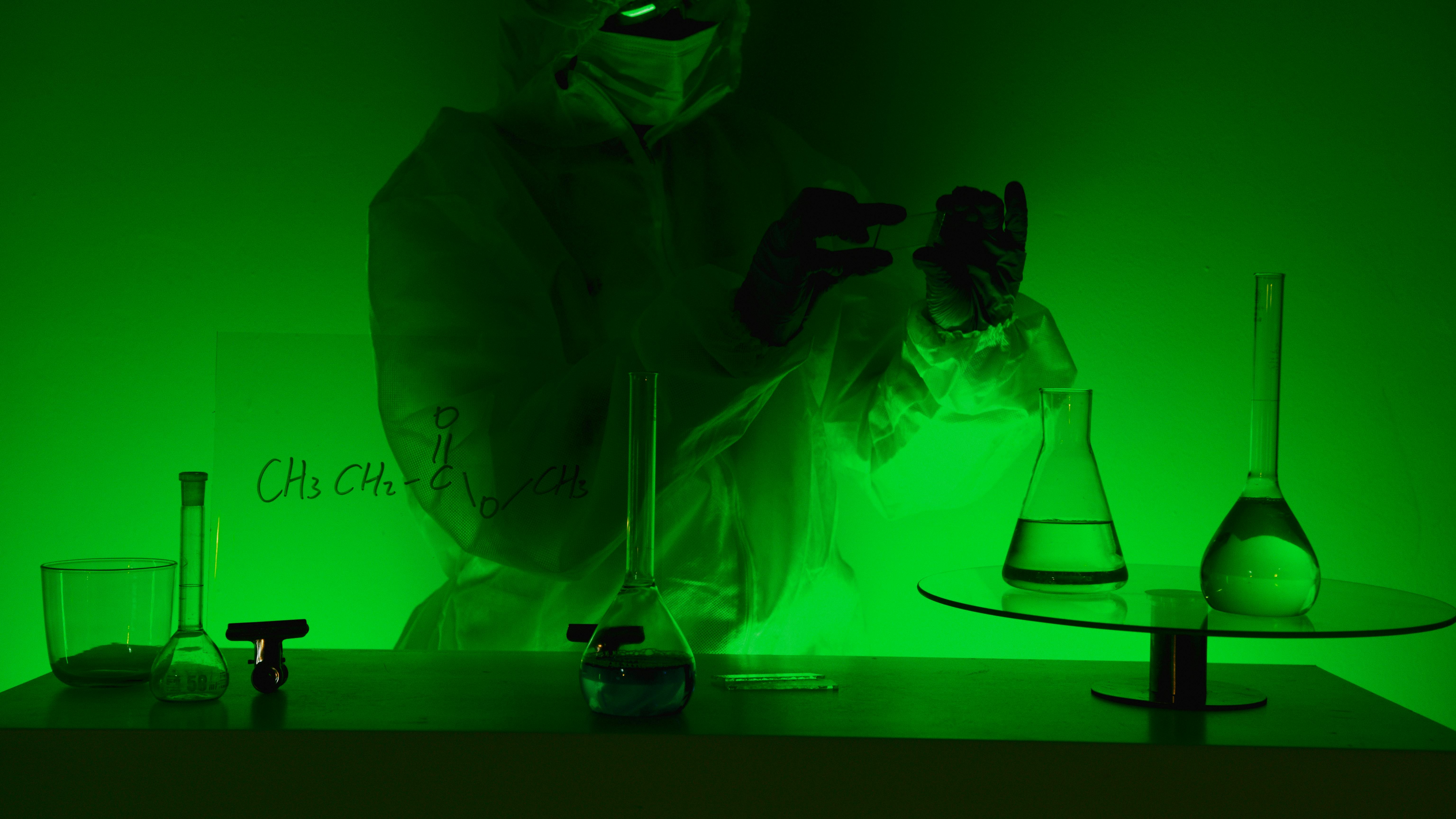 A scientist in protective gear works with glassware in a green-lit laboratory, evoking mystery and scientific exploration.