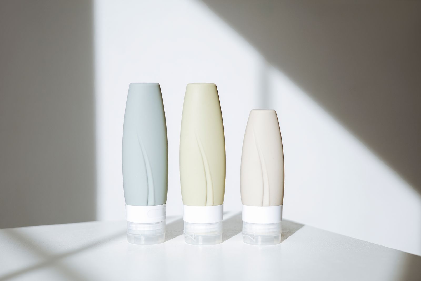 Three soft gray, sage and cream skincare bottles on a white table in angled sunlight and shadow