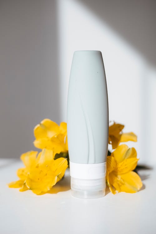 Close-Up Shot of a Lotion Bottle and Yellow Flowers on White Surface