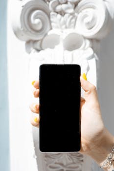 A close-up of a smartphone held by a hand against a decorative white background in bright light.