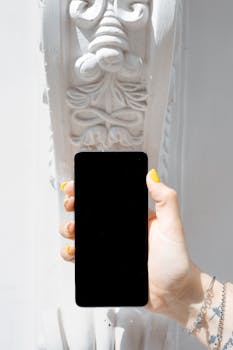 Hand with yellow nails holding smartphone against detailed carved white wall.