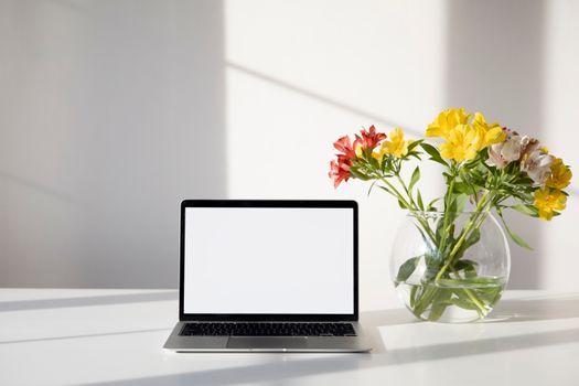 A sleek laptop with a blank screen next to a vibrant flower vase on a sunlit table.