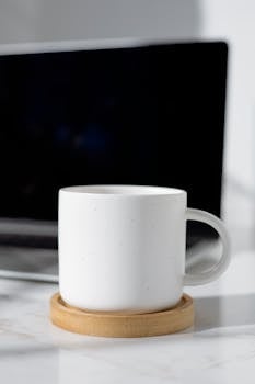 Simple white ceramic mug placed on a wooden coaster with a laptop in the background, ideal for workspace design.