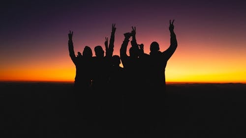 Figures of anonymous friends raising hands up while cheering and standing against colorful orange sundown and dark purple sky in evening