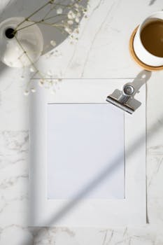 Minimalist flat lay with blank paper and binder clip on marble surface, beside a cup of coffee and a flower vase.