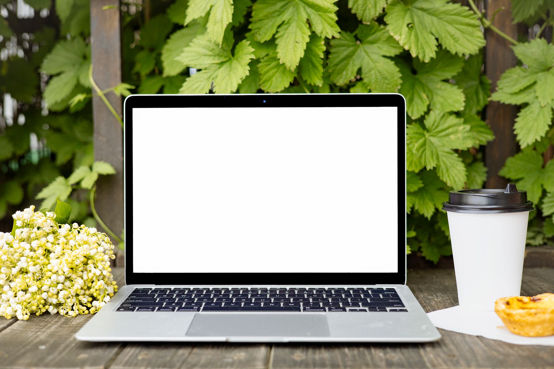 Open laptop with blank screen, coffee cup, and flowers on a wooden table outdoors.