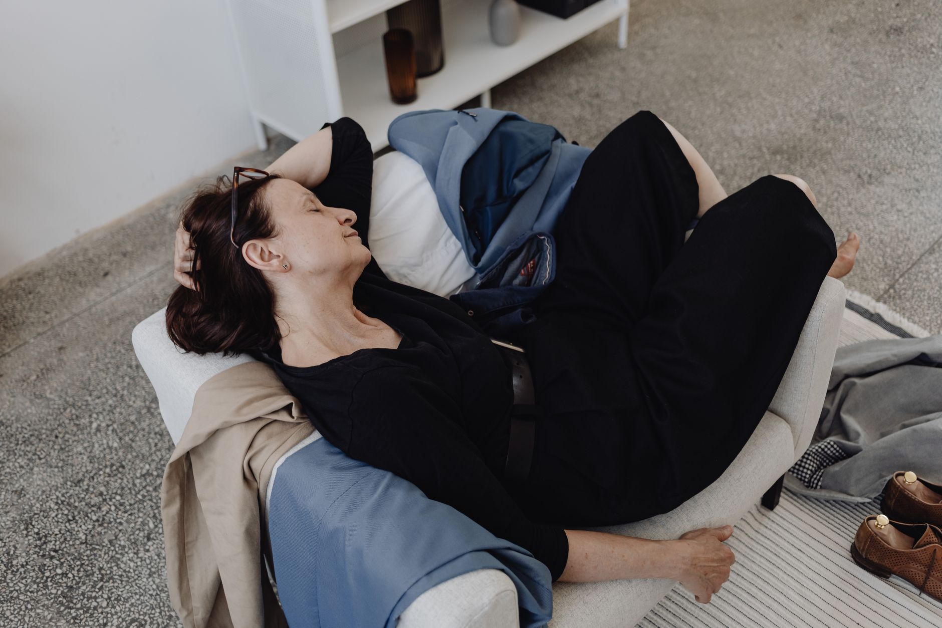A woman rests on a sofa indoors, surrounded by casual clothing and shoes.