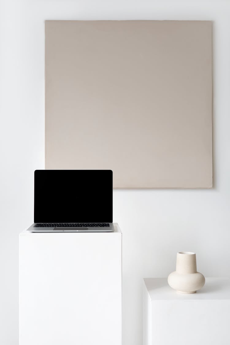 A Laptop On A White Platform Beside A Ceramic Vase