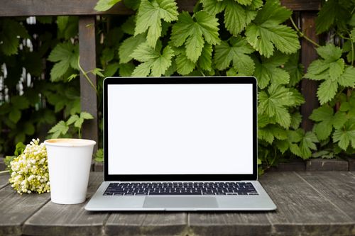 Free Laptop on wooden table with foliage backdrop and coffee cup, perfect for mockup use. Stock Photo