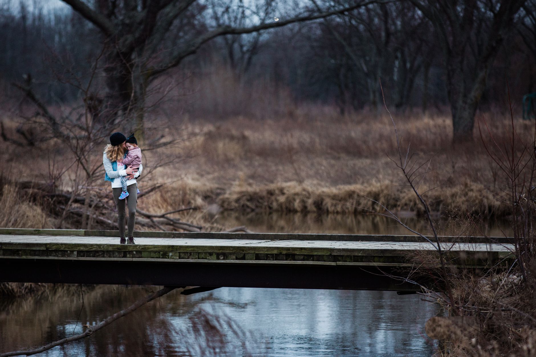 A mother holding her child on a bridge in a serene forest landscape.