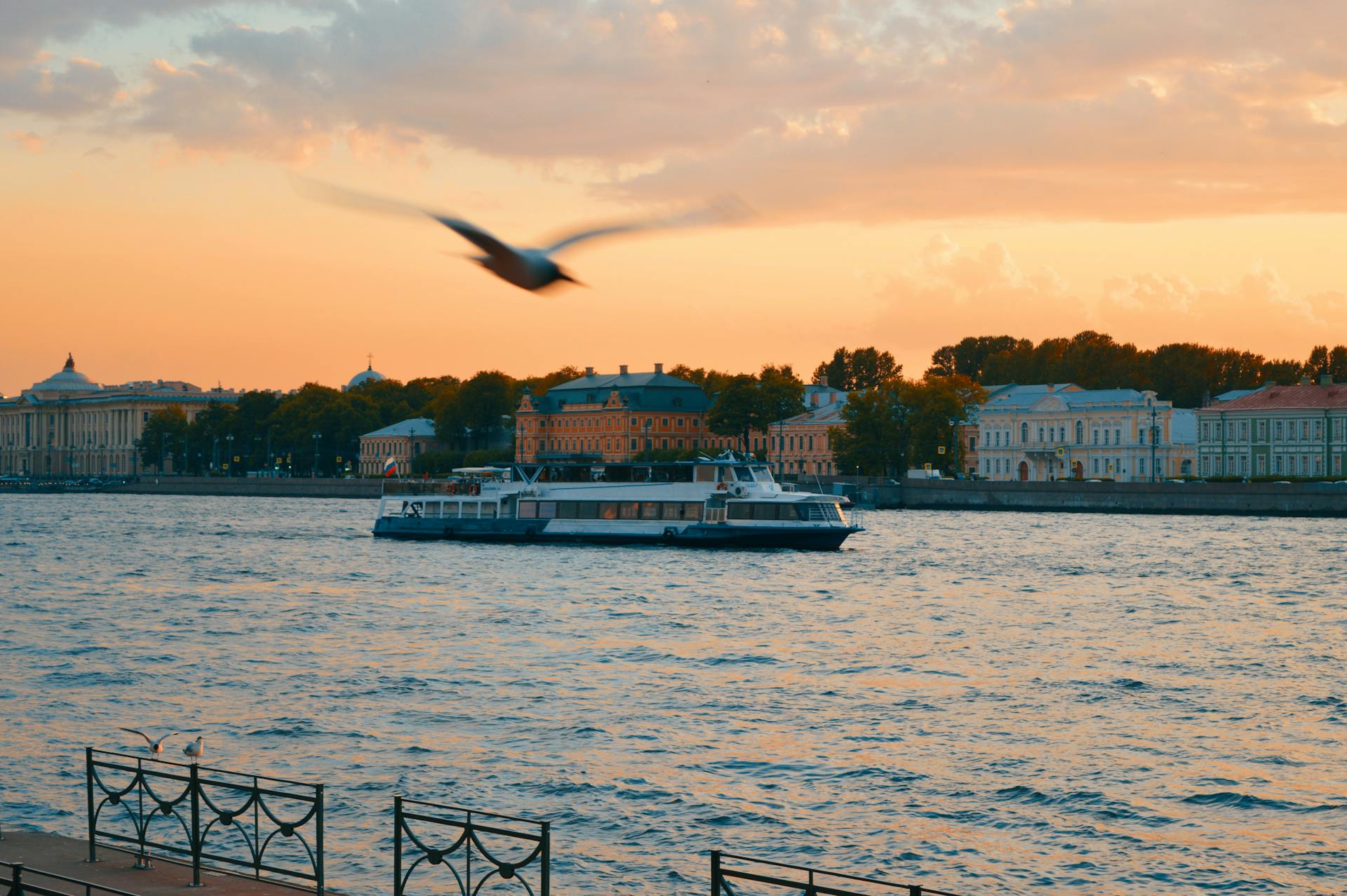 A peaceful ferry boat cruises along the river at sunset with a bird flying by.