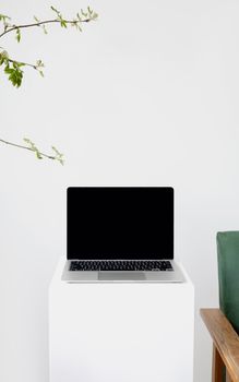 Sleek laptop setup on a pedestal with branches and chair in a minimalist white setting.