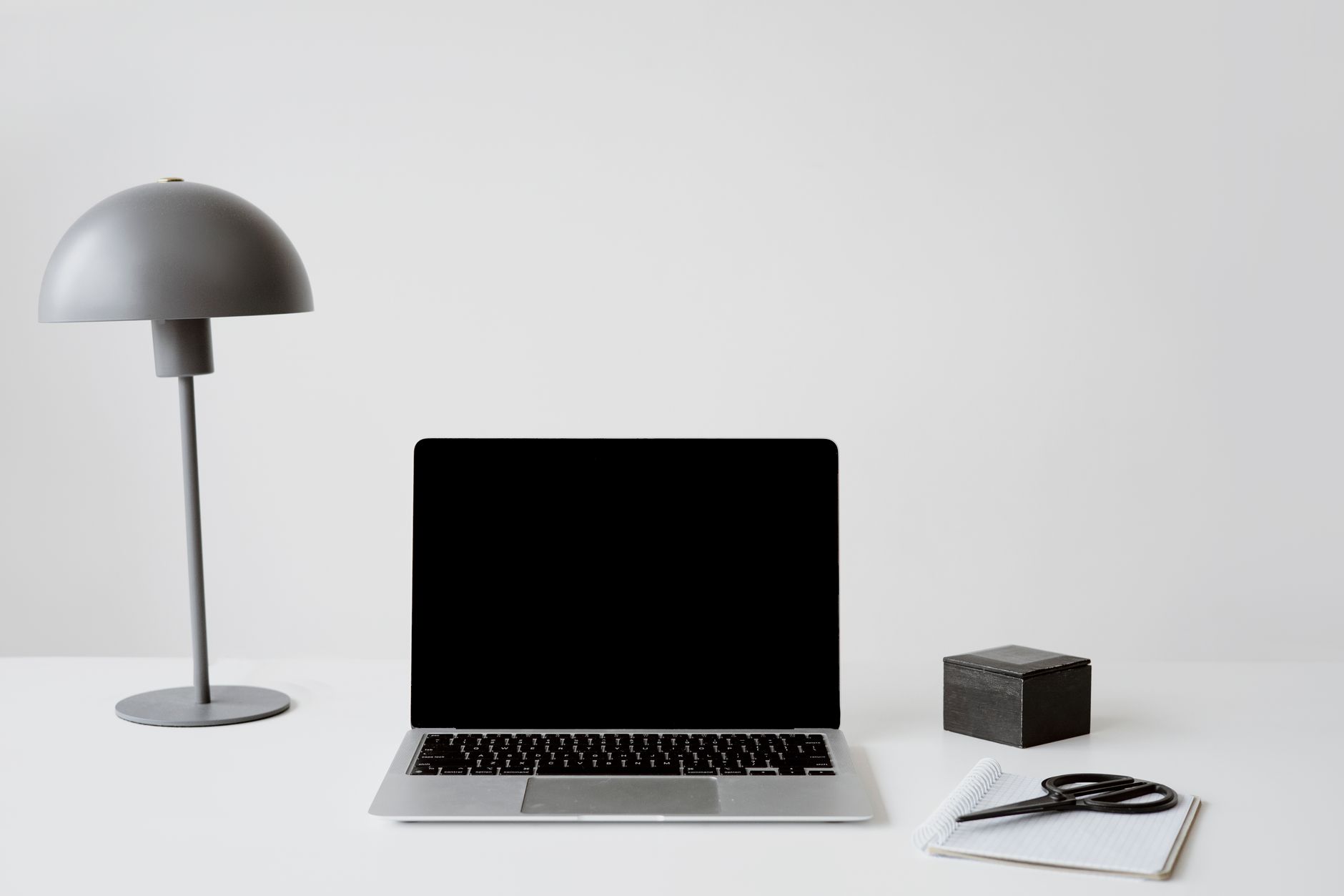 A minimalist office setup featuring a closed laptop, modern lamp, and a notepad with scissors on a white background.