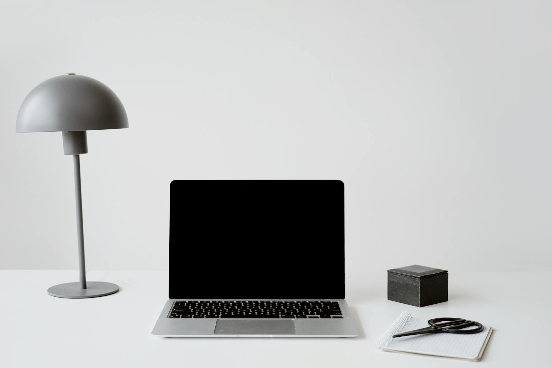 A minimalist office setup featuring a closed laptop, modern lamp, and a notepad with scissors on a white background.