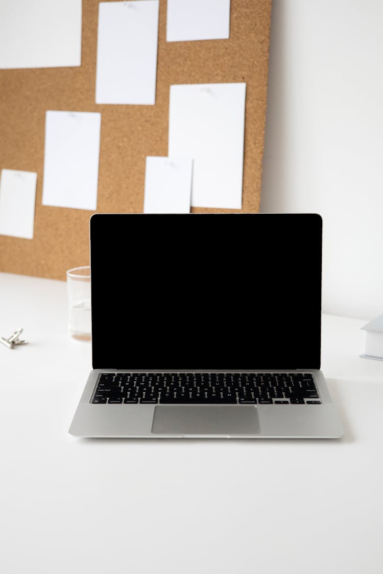 A Laptop Beside A Corkboard On A White Desk