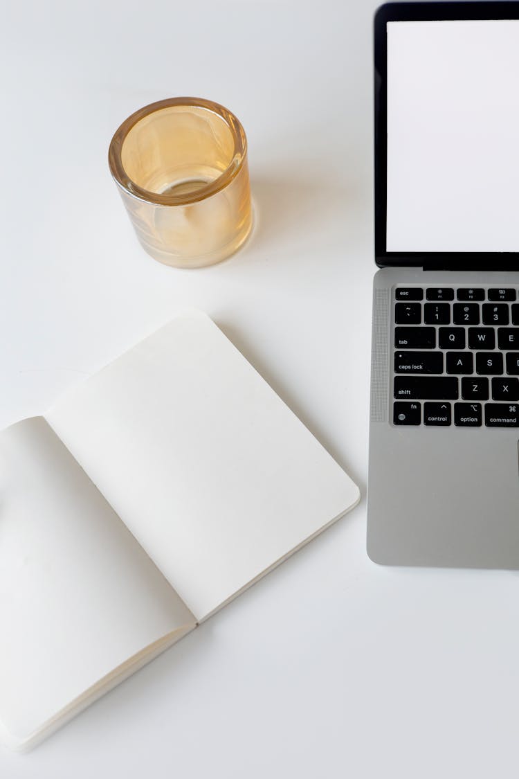 A Blank Notebook Beside A Laptop On A White Surface