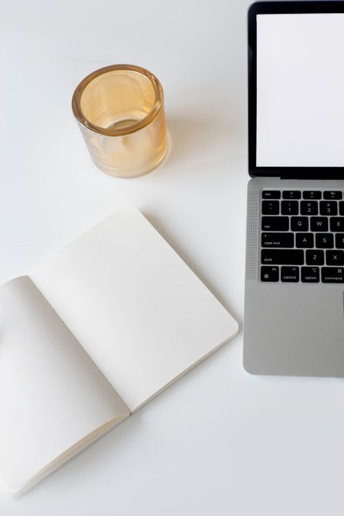Free Clean and minimalist workspace featuring an open notebook, laptop, and a glass cup on a white surface. Stock Photo