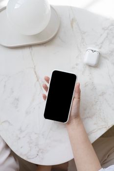 A person holding a smartphone on a marble table with AirPods nearby, ideal for tech and lifestyle themes.