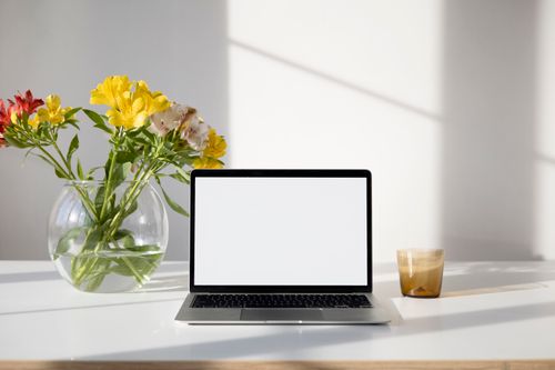 Free Minimalistic desk setup featuring a laptop, vase of flowers, and a cup under natural light. Stock Photo
