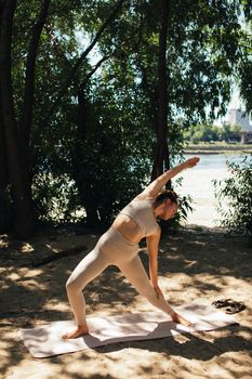 Woman practicing yoga on a mat under trees by water, embracing mindfulness and wellness.