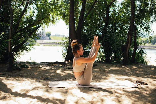 A woman practices yoga on a sandy surface under the trees near a river, enjoying a peaceful outdoor environment.