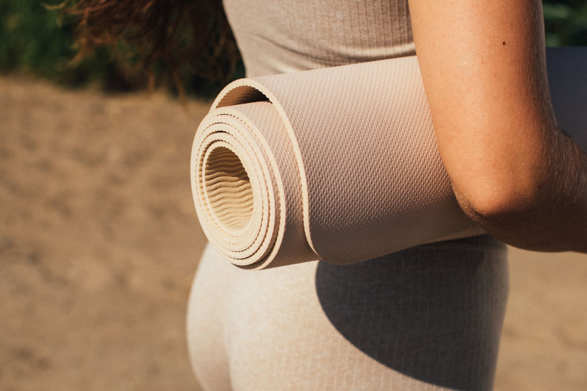 瑜珈墊材質比較:PVC、TPE、橡膠、棉質哪種最適合你? Person holding a rolled yoga mat in a sunlit outdoor setting, emphasizing fitness and wellness.