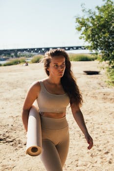 Fit woman walks on a sunny beach carrying a yoga mat, ready for outdoor exercise.