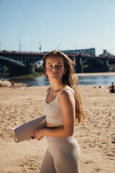Woman holding a yoga mat at the beach with a cityscape backdrop. Perfect for fitness and lifestyle themes.