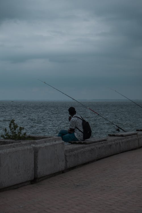 Man Sitting on a Pier with Two Fishing Rods Next to Him 