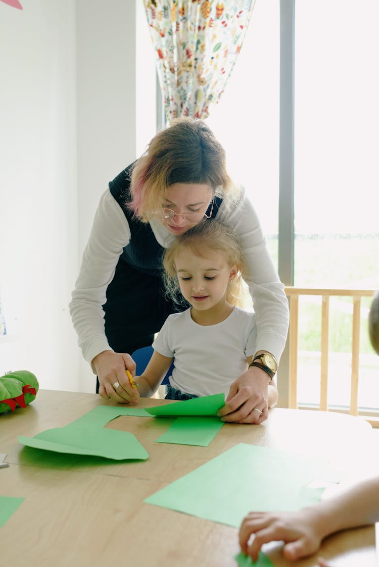 A Girl Sitting At The Table While Cutting Green Paper