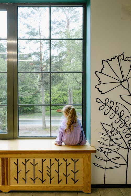 Free A young girl looks out a large window, surrounded by artistic wall decor. Stock Photo