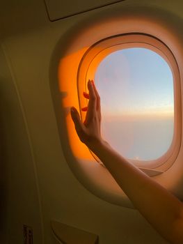 A hand reaches out to the warm golden sunset sky through an airplane window.
