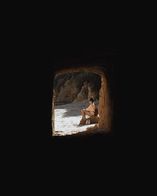 A woman sits peacefully in a cave entrance in the Faro District, Portugal, framed by rocky formations.