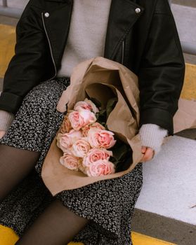 A woman in a floral dress holds a bouquet of pink roses wrapped in parchment paper indoors.