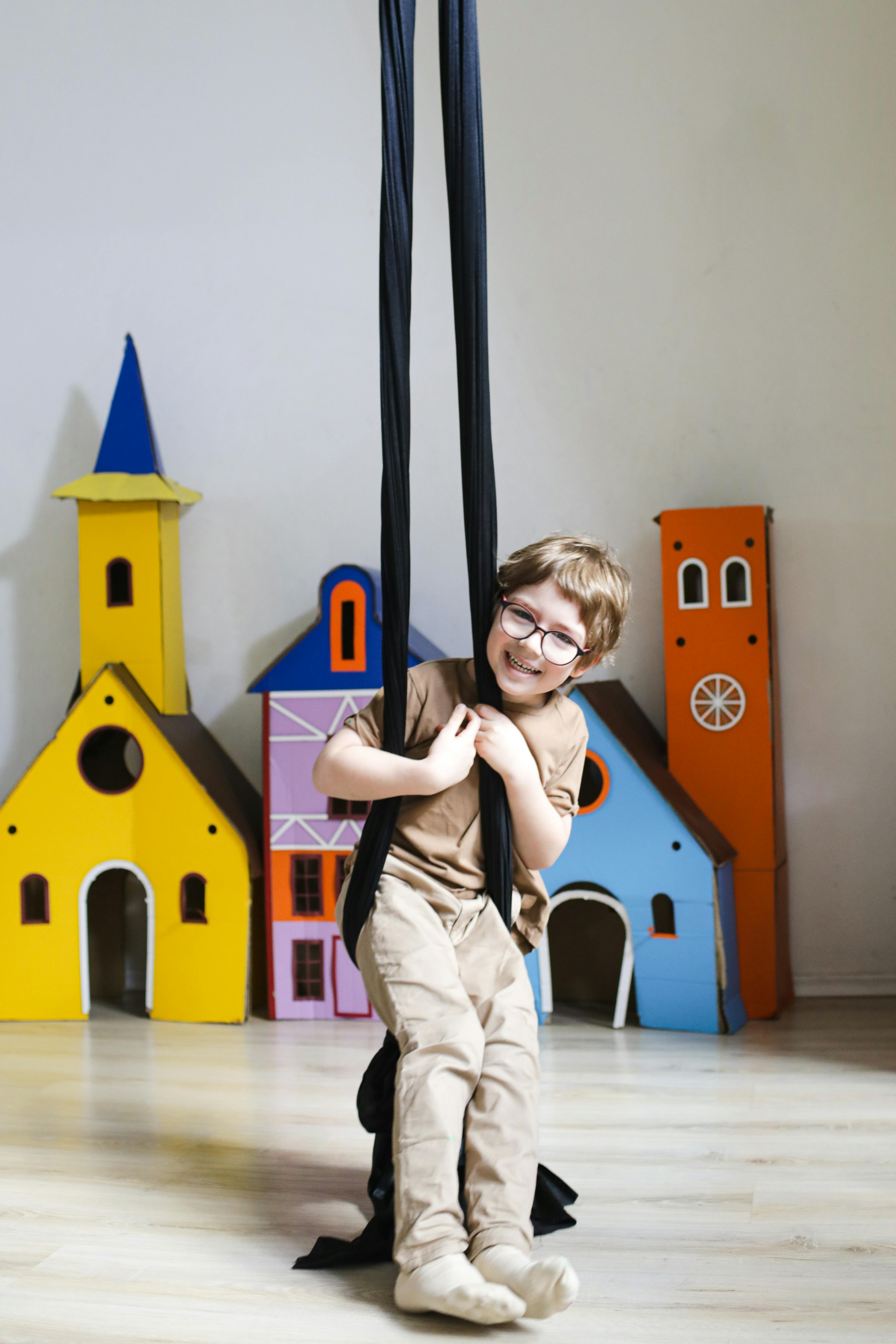 Free Joyful child with glasses swinging indoors with colorful playhouses in the background. Stock Photo