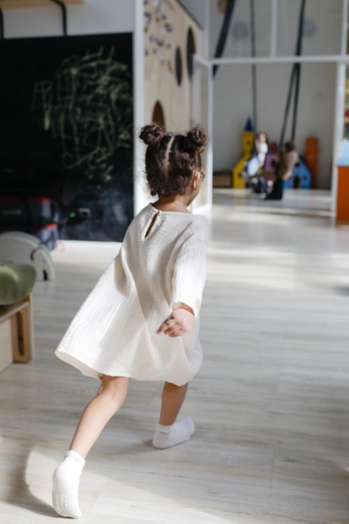 Free A young girl with hair buns runs joyfully in a sunlit playroom wearing a white dress. Stock Photo