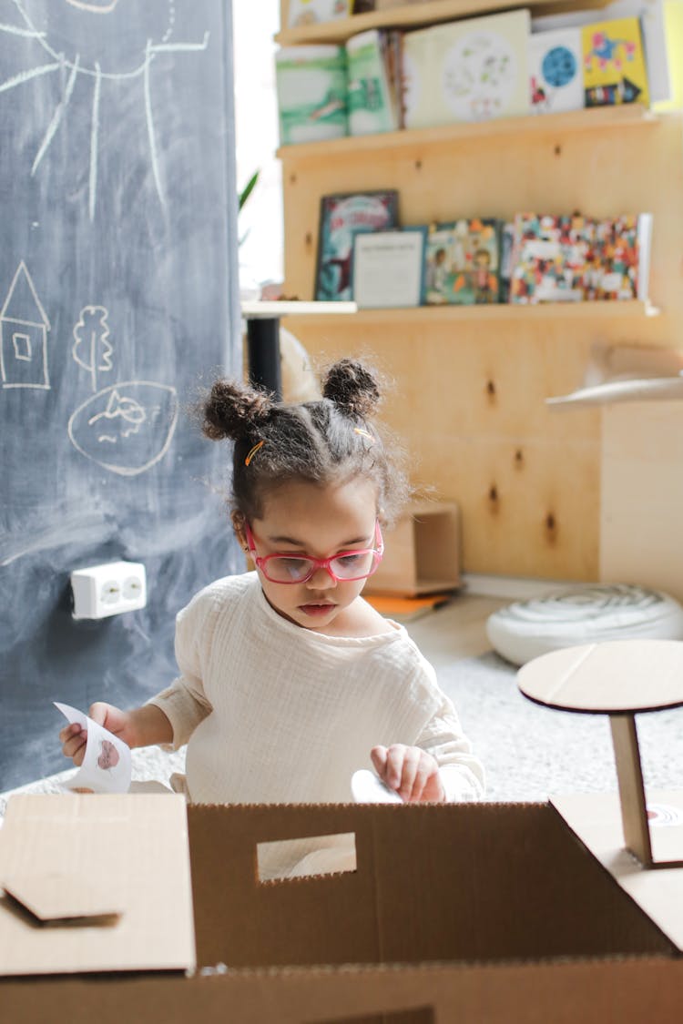 A Child In White Long Sleeves Holding Paper Stickers On A Carboard Box