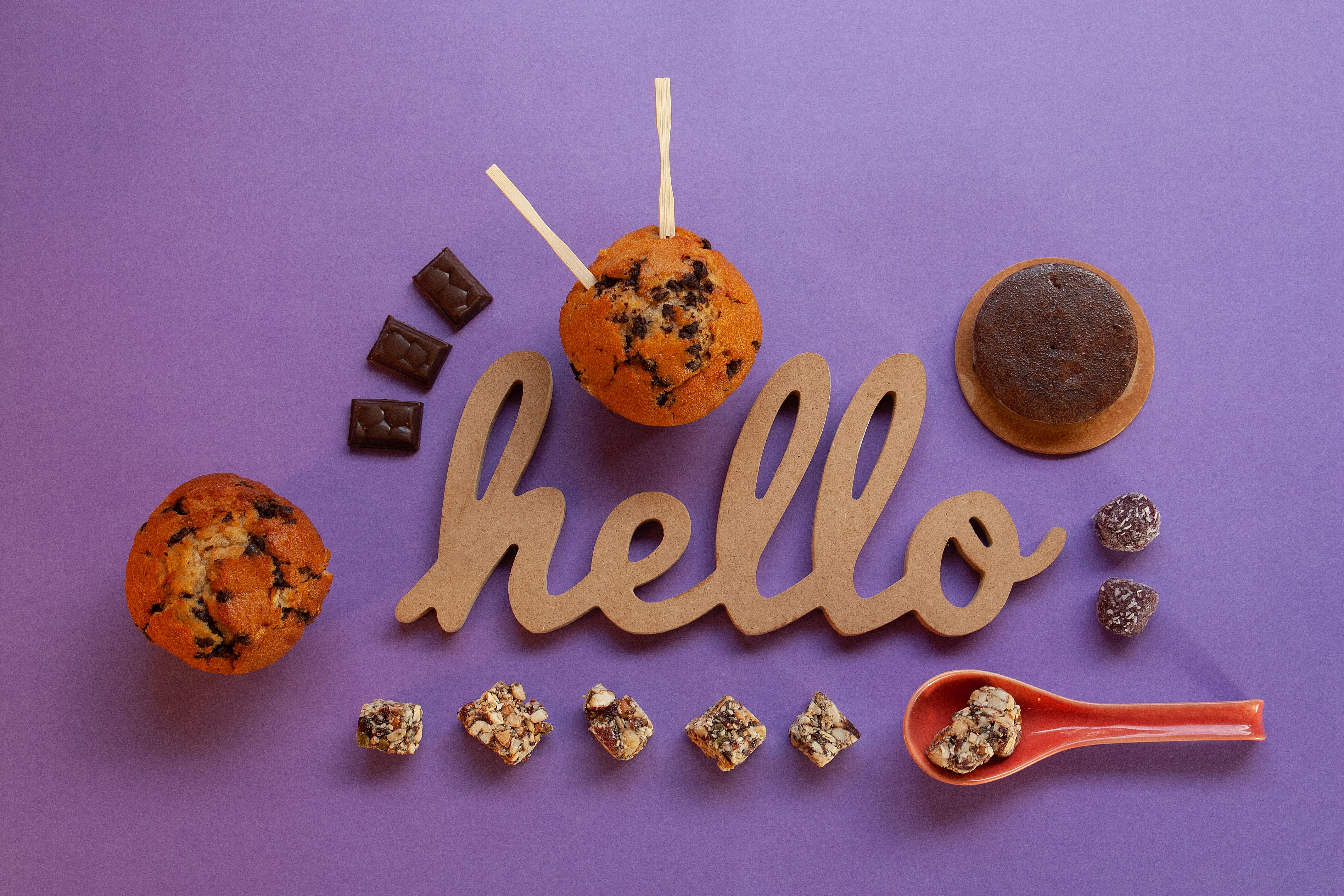 A top view of assorted chocolates and muffins with a 'Hello' sign on a purple surface.