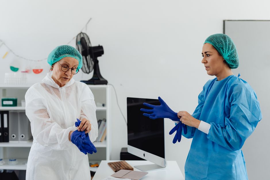 Dos mujeres trabajadoras de la salud en vestimenta quirúrgica poniéndose guantes de protección en una clínica.